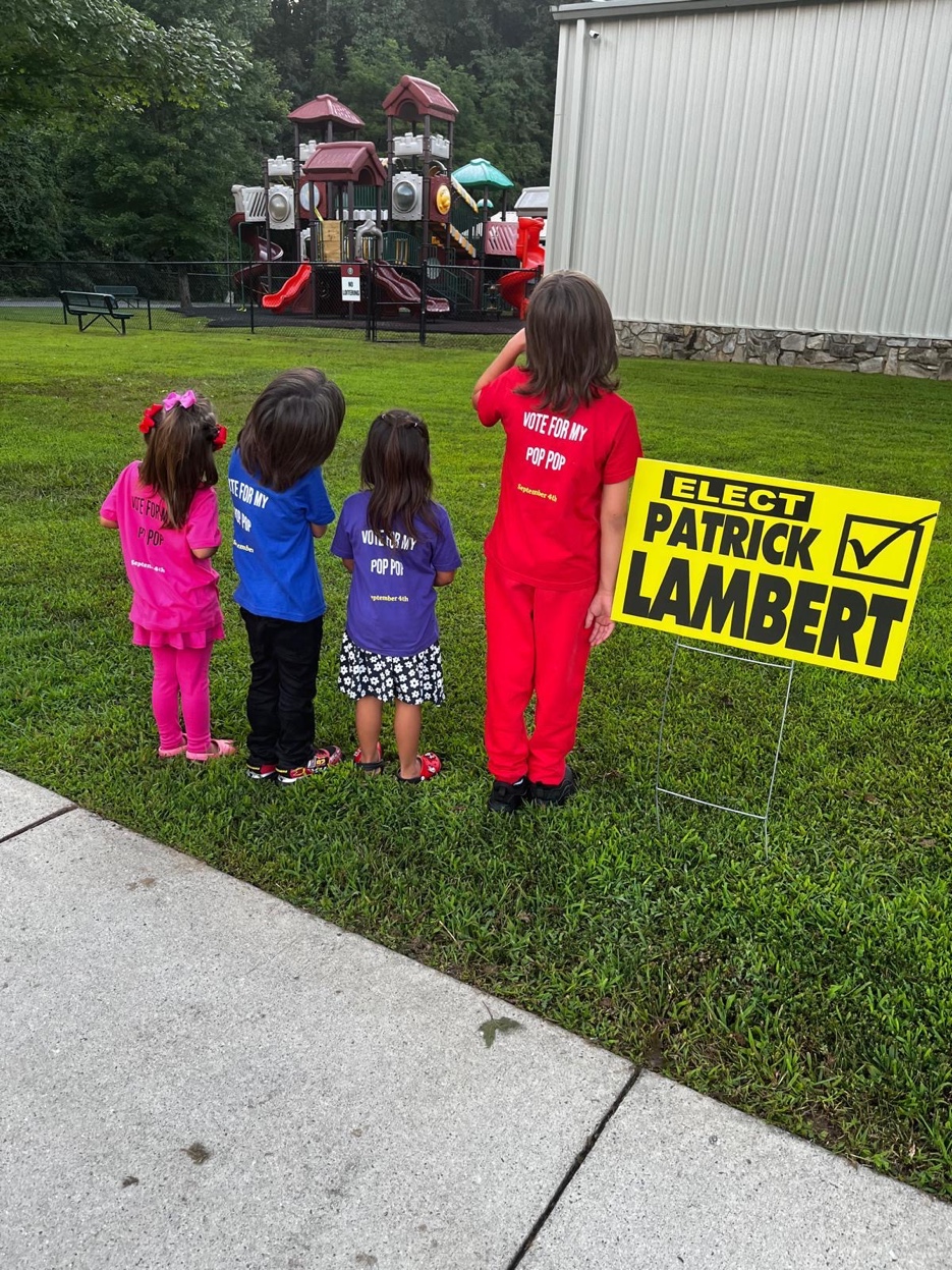 Four grandchildren in Vote For My Pop Pop t-shirts beside an Elect Patrick Lambert yard sign, 2015